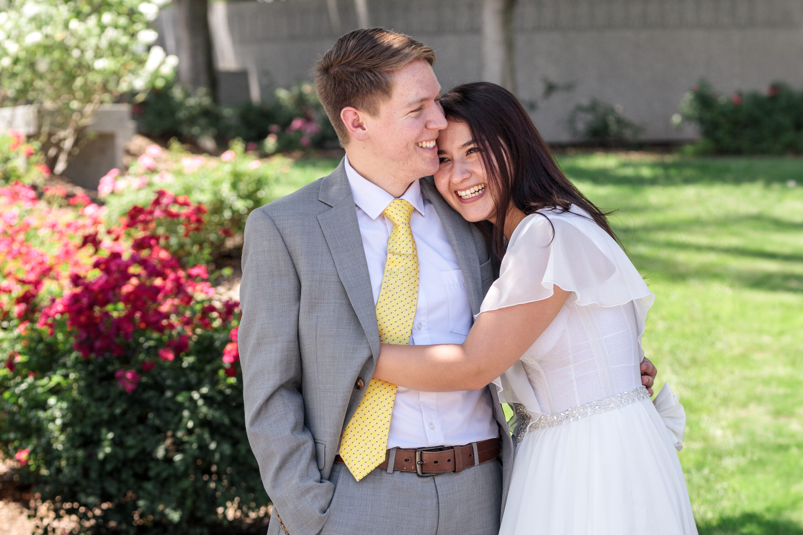 Happy couple on their wedding day hugging with big smiles on the beautiful grass and flowered grounds of the Redlands LDS Temple