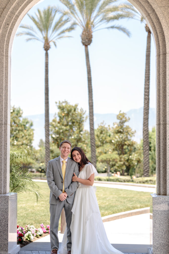 Couple smiling in the arch of the Redlands California LDS temple with palm trees behind them. 